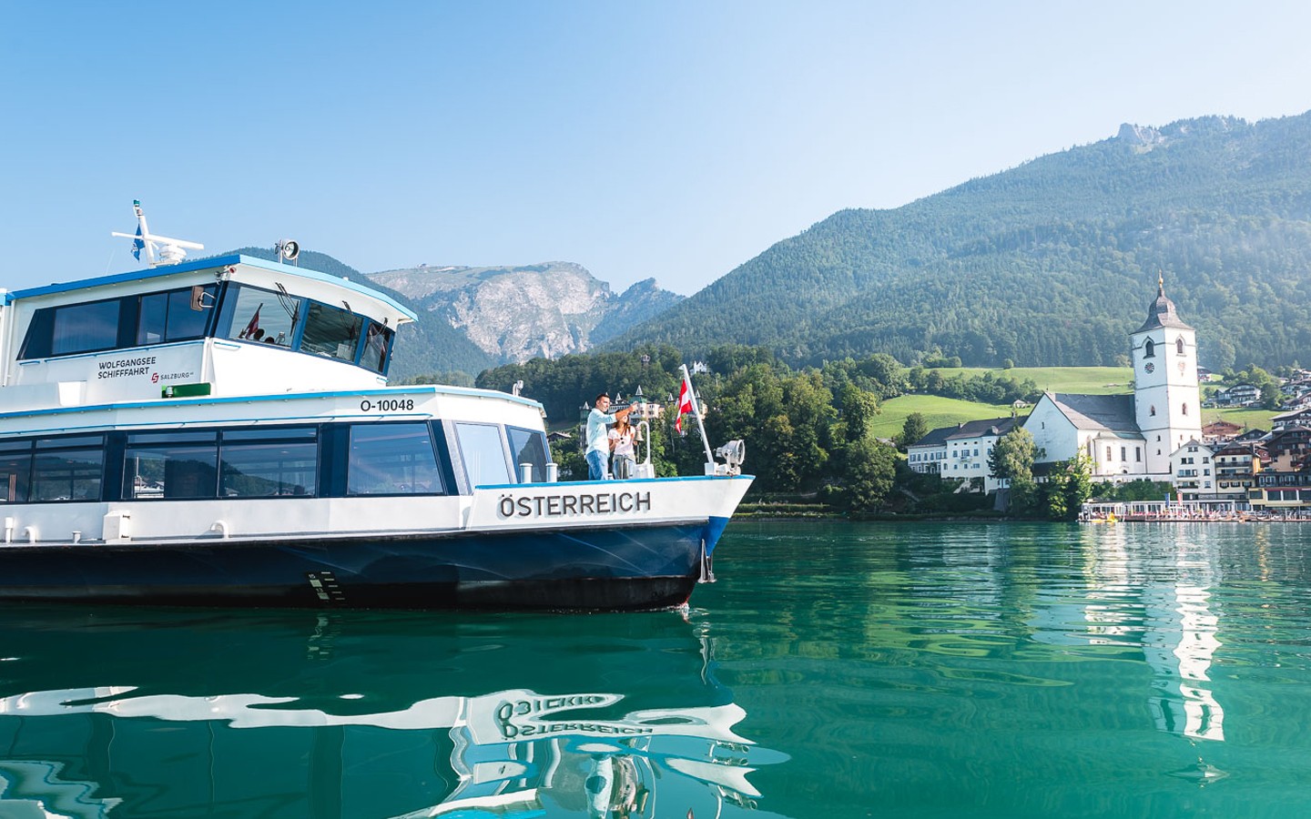 Zwei Personen stehen am Deck des Schiffes MS Salzburg im Hintergrund sieht man die Kirche von St. Wolfgang