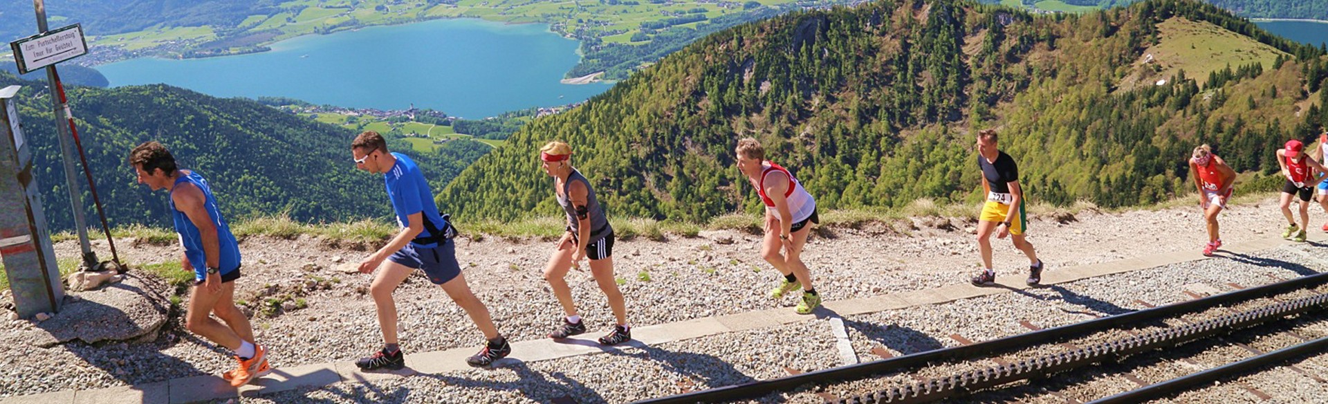 Personen laufen den Schafberg neben der Gleisanlage rauf. Im Hintergrund sieht man den Wolfgangsee
