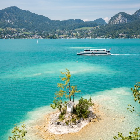 Ein Schiff der WolfgangseeSchifffahrt auf dem Wolfgangsee. Im Vordergrund sieht man die kleine Insel mit dem Ochsenkreuz drauf