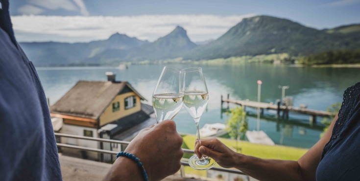 Zwei Personen stoßen mit Weingläsern auf der Seeterrasse des Café & Restaurant EQ an. Im Hintergrund sind man den Wolfgangsee und eine beeindruckende Berglandschaft im Sommer 