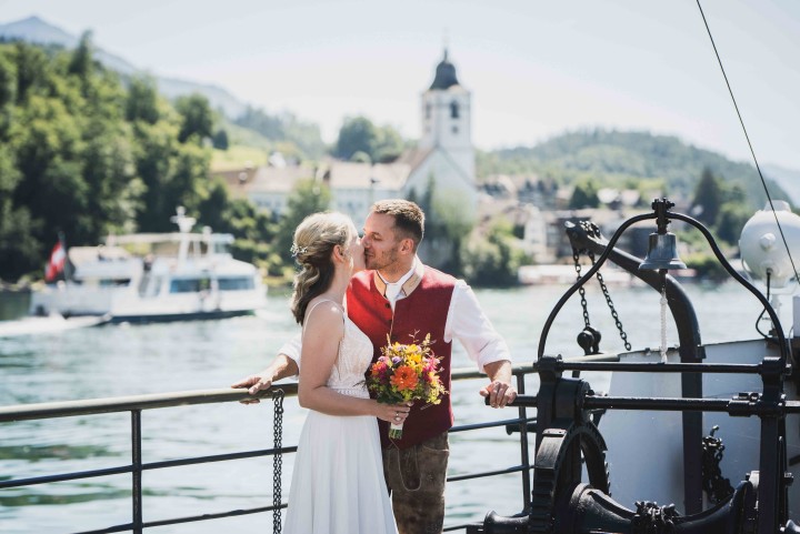 Hochzeitspaar küsst sich auf dem Schiff mit Blick vom Wolfgangsee aus auf die Kirche von St. Wolfgang