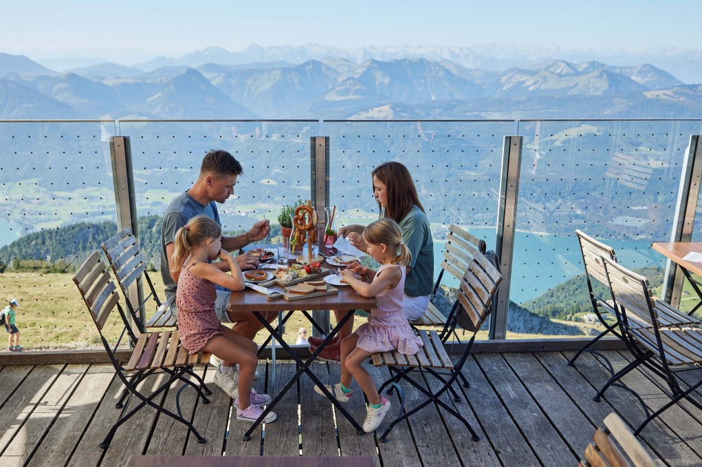 Eine Familie sitzt am Tisch auf der Terrasse der Himmelspforte am Schafberg mit Blick auf Wolfgangsee und Berge